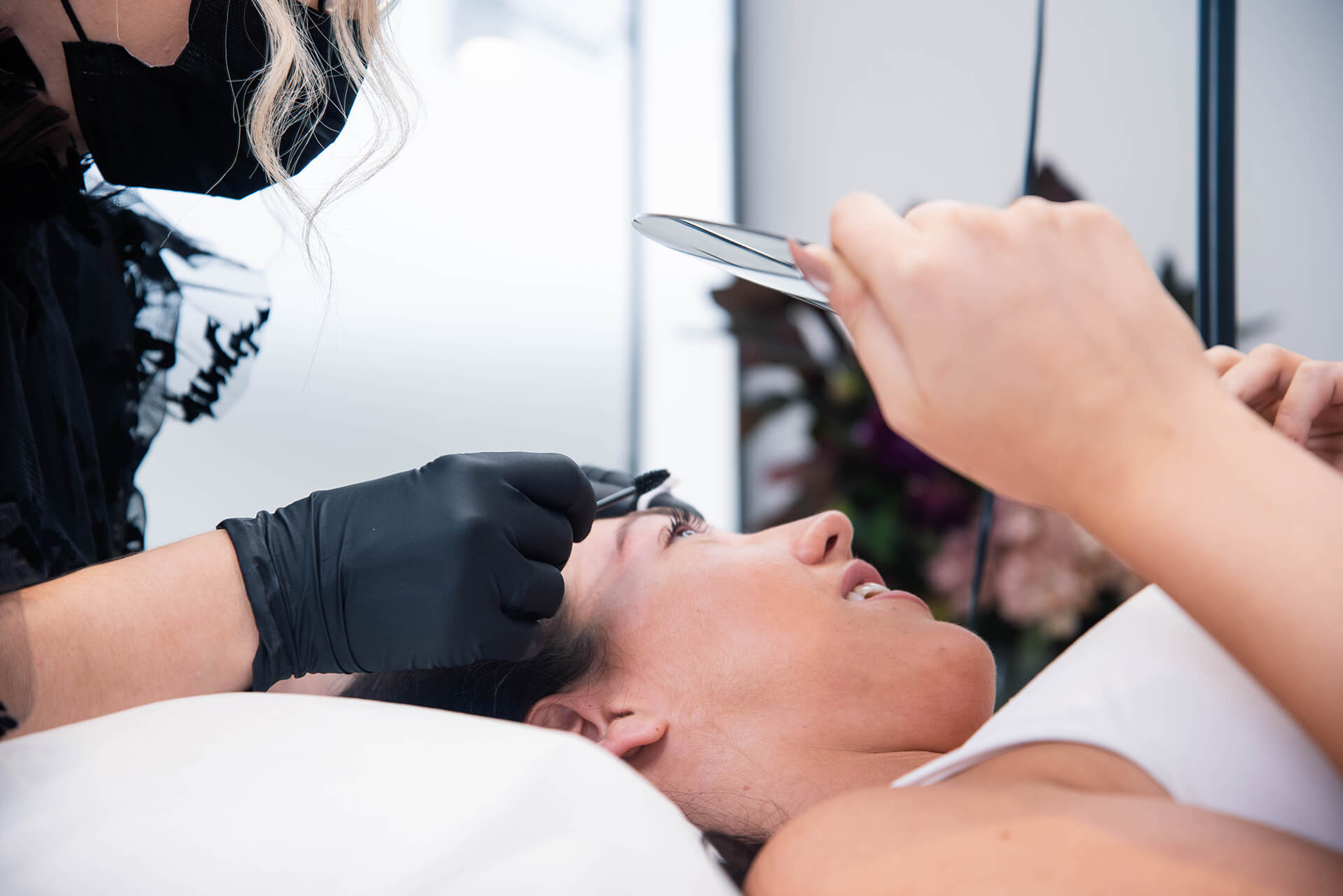 client lying down, holding a mirror, while our brow specialist performs precise eyebrow shaping